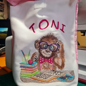 May include: White fabric lunch bag with a pink top and trim. Embroidered image of a monkey wearing glasses and a bow tie, with the name "TONI" above. The monkey is sitting at a desk with books and pencils. A small label at the bottom reads "Mit liebe gemacht".