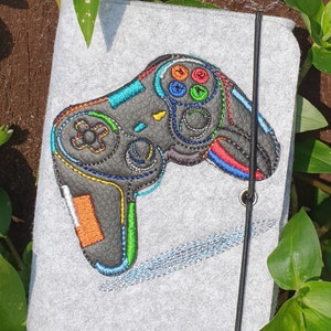 Könnte beinhalten: Eine graue Filz-Tasche mit einem schwarzen Gummibandverschluss. Die Tasche zeigt eine gestickte bunte Videospiel-Steuerung.