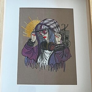 Op de afbeelding: Geborduurd kunstwerk in een houten lijst. Het kunstwerk toont een persoon met een paarse hoodie, zonnebril en gasmasker. De persoon heeft blauw haar en rode lippenstift. Een geel zonachtig ontwerp staat op de achtergrond.