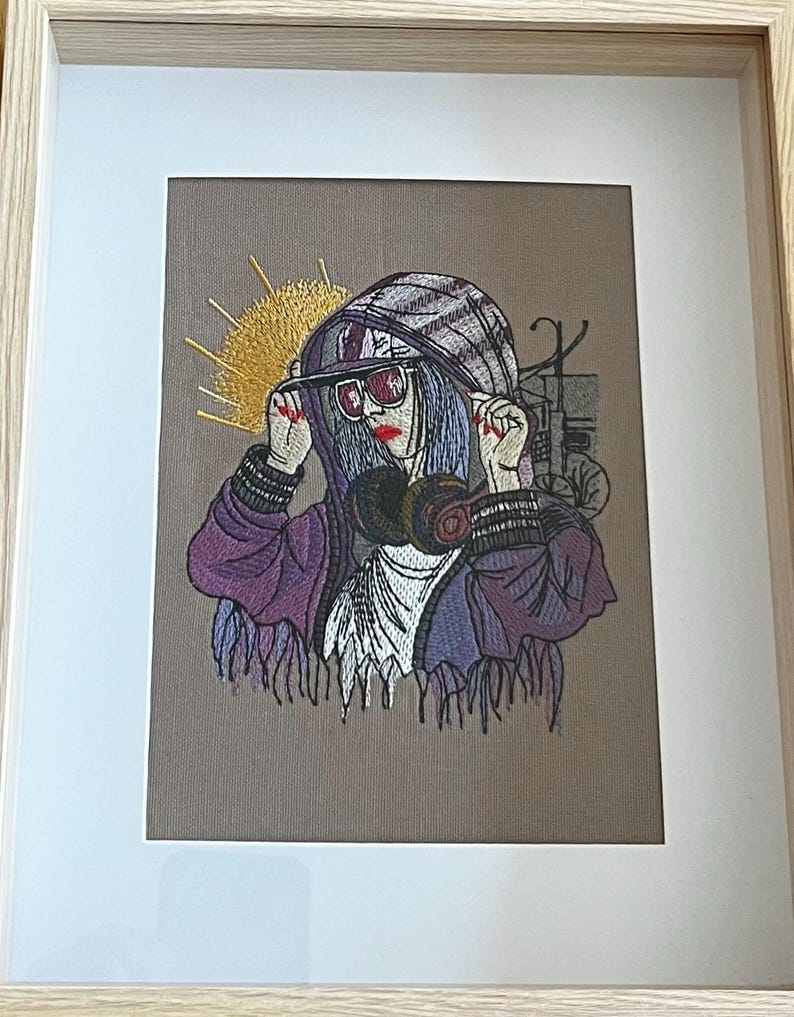 Op de afbeelding: Geborduurd kunstwerk in een houten lijst. Het kunstwerk toont een persoon met een paarse hoodie, zonnebril en gasmasker. De persoon heeft blauw haar en rode lippenstift. Een geel zonachtig ontwerp staat op de achtergrond.