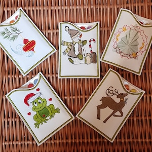 May include: Five white cardstock gift card holders with green trim and different embroidered Christmas designs. The designs include a snowman, a reindeer, a frog wearing a Santa hat, a Christmas tree, and a Christmas ornament.