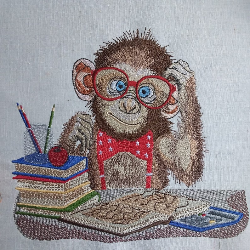 May include: Embroidered image of a monkey wearing red glasses and a bow tie with white stars. The monkey is sitting at a desk with books, pencils, and a calculator. A red apple sits atop the books.