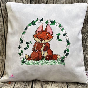 May include: A white pillow with an embroidered design of a red fox sitting in a circle of green leaves. The fox has its eyes closed and is holding its paws together.