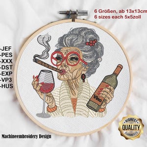 Op de afbeelding: Geborduurd ontwerp in een houten hoepel met een vrouw met grijs haar, een rode bril en een sigaar, die een wijnglas en een fles met het opschrift "Vino" vasthoudt. Het ontwerp bevat tekst en een gouden "Highest Quality Guaranteed" zegel.