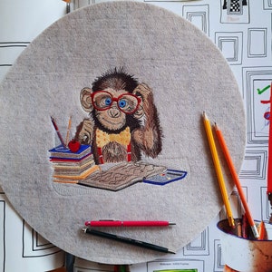 May include: Embroidered felt circle featuring a monkey wearing red glasses and a yellow bow tie, sitting at a desk with books and pencils. The monkey is brown with blue eyes. Pens and pencils are scattered around the felt circle.