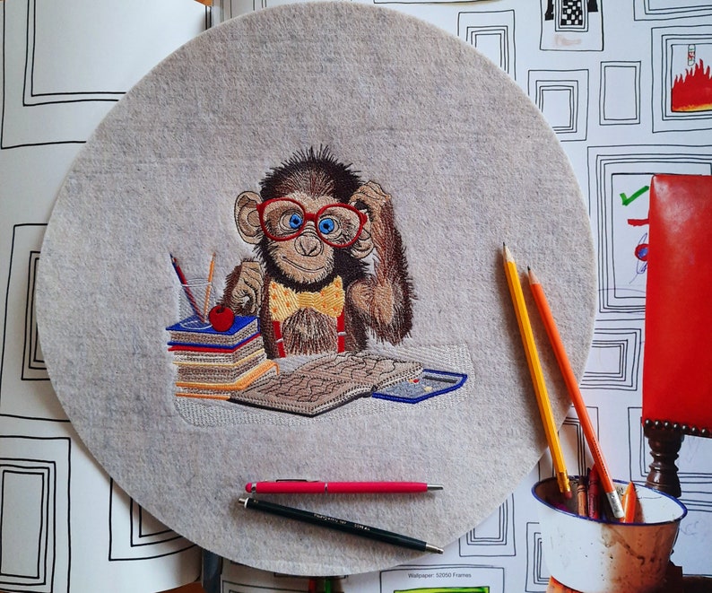May include: Embroidered felt circle featuring a monkey wearing red glasses and a yellow bow tie, sitting at a desk with books and pencils. The monkey is brown with blue eyes. Pens and pencils are scattered around the felt circle.