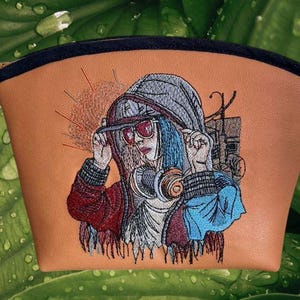 Op de afbeelding: Een bruin make-up tasje met een zwarte rits en een gedetailleerd geborduurd ontwerp. Het ontwerp toont een persoon met een hoodie, zonnebril en koptelefoon. De persoon heeft blauw haar. De achtergrond is een groen blad.