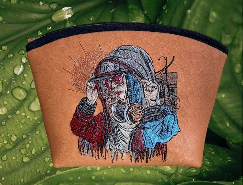 Op de afbeelding: Een bruin make-up tasje met een zwarte rits en een gedetailleerd geborduurd ontwerp. Het ontwerp toont een persoon met een hoodie, zonnebril en koptelefoon. De persoon heeft blauw haar. De achtergrond is een groen blad.