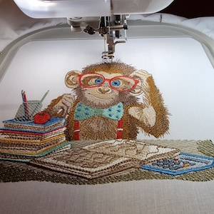 May include: An embroidered design of a brown monkey wearing red glasses and a blue bow tie, seated at a desk. The monkey is surrounded by books, pencils, and a calculator. The design is being created by an embroidery machine.