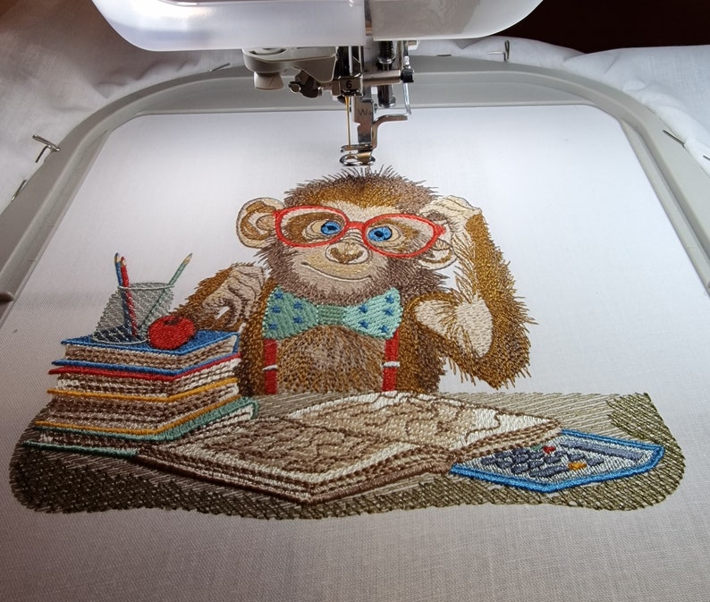 May include: An embroidered design of a brown monkey wearing red glasses and a blue bow tie, seated at a desk. The monkey is surrounded by books, pencils, and a calculator. The design is being created by an embroidery machine.
