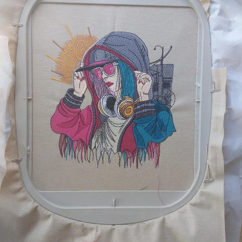 Op de afbeelding: Geborduurd ontwerp van een persoon met een hoodie, zonnebril en koptelefoon. Het ontwerp heeft een kleurenpalet van roze, blauw en grijs, met een zonnestraalontwerp op de achtergrond. De borduursel is in een borduurring.