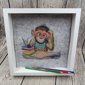 May include: Embroidered artwork of a monkey wearing red glasses and a blue bow tie, framed in white. The monkey is sitting at a desk with books, pencils, and a calculator. Three colored pencils rest in front of the frame.