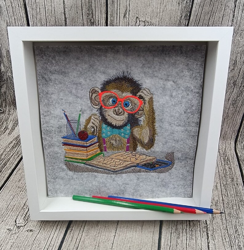 May include: Embroidered artwork of a monkey wearing red glasses and a blue bow tie, framed in white. The monkey is sitting at a desk with books, pencils, and a calculator. Three colored pencils rest in front of the frame.