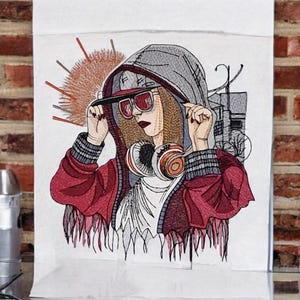 Op de afbeelding: Geborduurd kunstwerk van een persoon met een rode jas, grijze hoodie en zonnebril, met een koptelefoon. Het kunstwerk heeft een streetstyle thema, met een stadsgezicht op de achtergrond en een zonnester ontwerp.