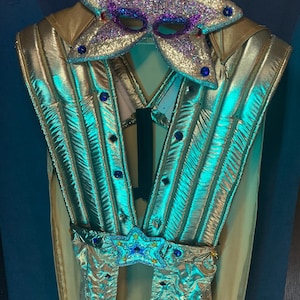 May include: A metallic gold and teal waistcoat featuring vertical quilted panels, adorned with dark blue circular gems. A matching star-shaped mask with purple glitter and blue gems is positioned at the top, and a star-shaped belt buckle is at the waist.