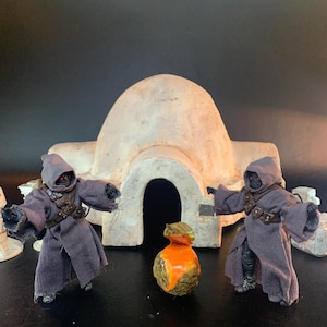 May include: Two action figures wearing dark gray hooded robes with brown utility belts stand on either side of a small orange and brown rock-like object. Behind them is a beige domed structure with a central arched doorway and two small white droids.