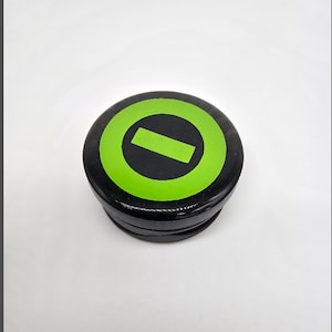 May include: A black, round object with a green circle and a diagonal black line. The object appears to be made of a hard material and has a slightly textured surface. The design is simple and modern.