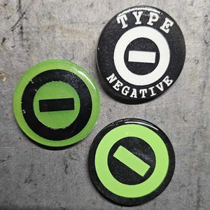 以下が含まれることがあります： 白の円と中央に黒い水平線がある、黒と緑の丸いボタンが3つ。 上のボタンには、円の周りに白い文字で「TYPE NEGATIVE」と書いてあります。