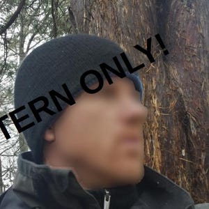 Op de afbeelding: Een persoon draagt ​​een zwarte gebreide beanie met de tekst "PATTERN ONLY!" op de voorkant. De persoon staat voor een boom.