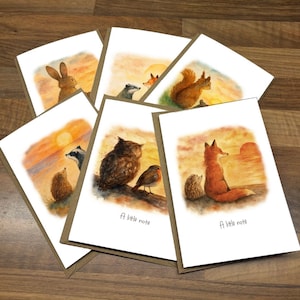 May include: A set of greeting cards featuring watercolour illustrations of woodland creatures such as a rabbit, fox, squirrel, owl, and hedgehog, set against a sunset. Each card has a brown border and the text "A little note".