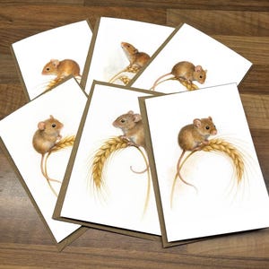 May include: A set of greeting cards with watercolour illustrations of brown mice on wheat. The cards have a white background and a brown border. They are arranged on a wooden surface.