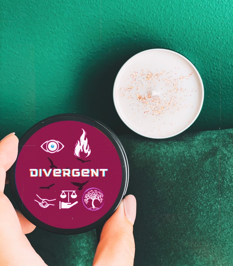 Puede incluir: Un tarro de vela rojo con la palabra "Divergent" y cinco s&iacute;mbolos: un ojo, una llama, una balanza, un &aacute;rbol y una mano con dos p&aacute;jaros volando por encima. El tarro de vela est&aacute; sobre una superficie verde.