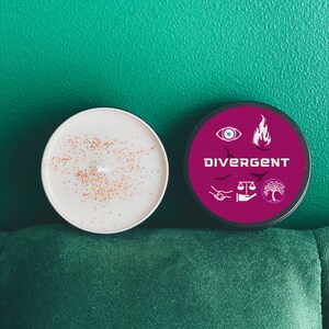 Puede incluir: Una vela blanca con purpurina dorada en la parte superior, junto a un tarro de vela negro con una etiqueta rosa que dice "Divergent" y tiene s&iacute;mbolos de una llama, un ojo, una balanza, un &aacute;rbol y dos manos que se dan la mano.