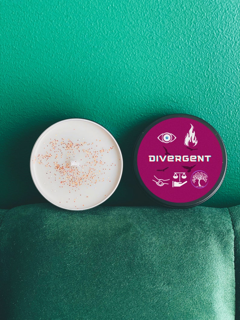 Puede incluir: Una vela blanca con purpurina dorada en la parte superior, junto a un tarro de vela negro con una etiqueta rosa que dice "Divergent" y tiene s&iacute;mbolos de una llama, un ojo, una balanza, un &aacute;rbol y dos manos que se dan la mano.