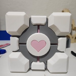 May include: A white and gray 3D printed cube with a pink heart in the center. The cube has a rounded, geometric design.