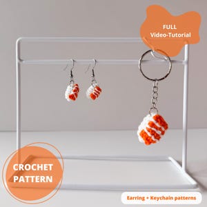 May include: A white metal display stand holds two crochet earrings and a keychain charm. The earrings and charm are white and orange, resembling sushi. The image includes text that says "CROCHET PATTERN" and "FULL Video-Tutorial".