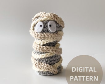 May include: A crocheted mummy toy with white eyes and a gray and beige striped body. The toy is sitting on a white surface with a brown circle that says "DIGITAL PATTERN" in white text.