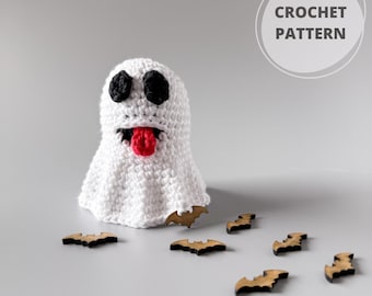 Puede incluir: Un fantasma blanco tejido a ganchillo con ojos negros y una lengua roja, rodeado de decoraciones de murciélagos de madera. La imagen incluye el texto "CROCHET PATTERN" en un círculo.