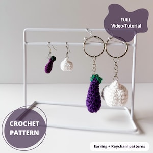 Puede incluir: Un expositor de metal blanco sostiene pendientes y llaveros de ganchillo. Los pendientes tienen forma de berenjena morada y diente de ajo blanco. Los llaveros presentan los mismos diseños. El texto "CROCHET PATTERN" y "FULL Video-Tutorial" son visibles.