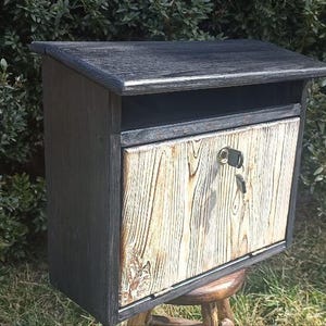 May include: A wooden mailbox with a black roof and frame, and a light-coloured, textured wooden door. The mailbox has a keyhole and is set against a backdrop of green foliage.
