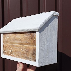Può includere: Una cassetta delle lettere in legno bianco con tetto spiovente e due cassetti. I cassetti hanno una finitura strutturata marrone chiaro. La cassetta delle lettere è tenuta contro uno sfondo di metallo ondulato marrone.