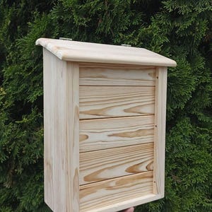 Może przedstawiać: Drewniana skrzynka pocztowa z naturalnym wykończeniem i poziomym usłojeniem drewna. Skrzynka pocztowa ma klapę na zawiasach i szczelinę na listy.