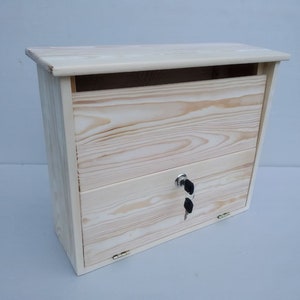 Könnte beinhalten: Ein Holzbriefkasten mit einer Klapptür und einem Schlüsselloch. Der Briefkasten ist aus hellem Holz gefertigt und hat eine natürliche Oberfläche.
