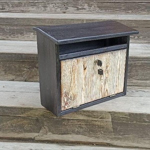 May include: A wall-mounted wooden mailbox featuring a dark charcoal frame and a light, natural wood grain front panel. The unit includes a top slot for mail and a hinged door with a central metal lock mechanism for secure access to contents.
