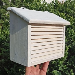 Può includere: Una cassetta postale da parete in legno bianco dotata di tetto spiovente e pannello frontale con lamelle orizzontali. La cassetta ha una forma rettangolare con una cerniera visibile sul bordo superiore e una finitura chiara e semplice.