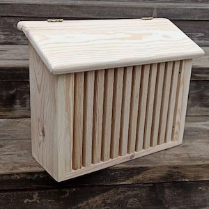 Może przedstawiać: Jasna drewniana skrzynka pocztowa z listwowym frontem i skośnym dachem. Skrzynka pocztowa wykonana jest z niewykończonego drewna i ma pokrywę na zawiasach. Skrzynka pocztowa stoi na drewnianej powierzchni.