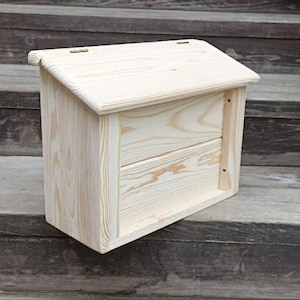 May include: A light-colored wooden mailbox with a sloped roof and a front panel. The mailbox is made of unfinished wood and sits on a set of wooden steps. The design features a hinged top for access.
