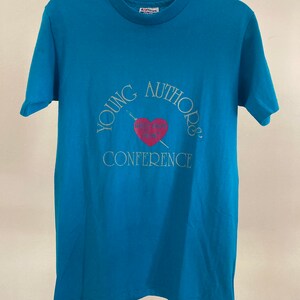 May include: A turquoise blue t-shirt with a white graphic that says "Young Authors Conference" with a heart and a pen.