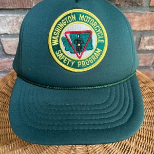 May include: Green trucker hat with a yellow and green embroidered patch that says "Washington Motorcycle Safety Program" and features a red triangle with a motorcycle and a person inside.