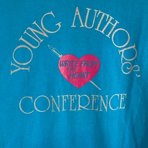 May include: A turquoise blue t-shirt with the text "YOUNG AUTHORS CONFERENCE" in silver glitter lettering. A pink heart with the text "WRITE FROM THE HEART" is in the center of the shirt.