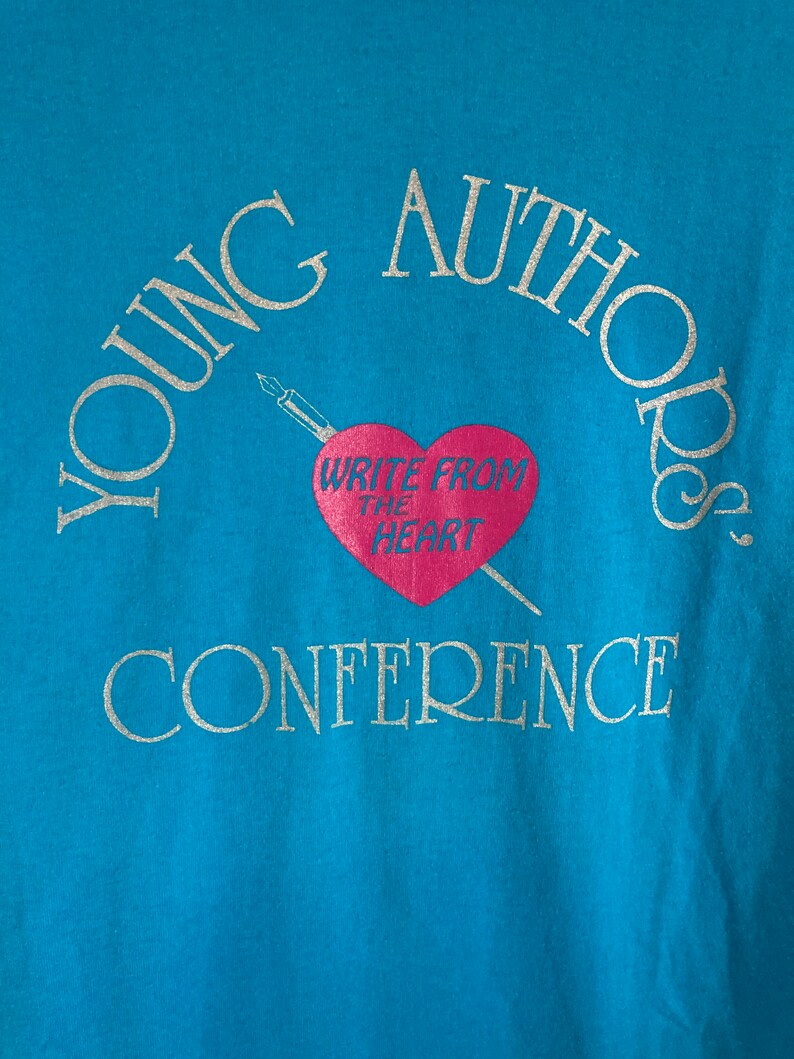 May include: A turquoise blue t-shirt with the text "YOUNG AUTHORS CONFERENCE" in silver glitter lettering. A pink heart with the text "WRITE FROM THE HEART" is in the center of the shirt.
