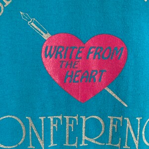 May include: A turquoise blue t-shirt with a silver graphic of a pen and heart. The heart is pink and says "Write from the heart". The bottom of the shirt says "Conference" in silver.