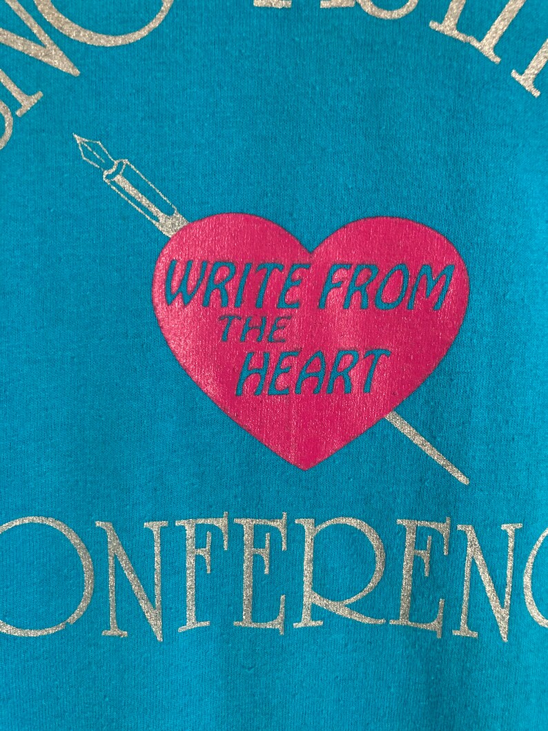 May include: A turquoise blue t-shirt with a silver graphic of a pen and heart. The heart is pink and says "Write from the heart". The bottom of the shirt says "Conference" in silver.