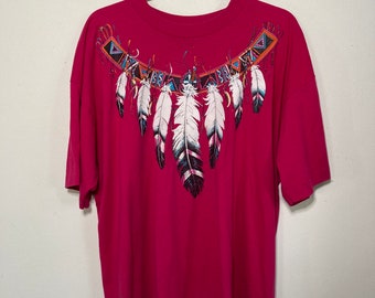 May include: A pink t-shirt with a graphic of a Native American inspired feather design. The design features a row of white feathers with black tips, surrounded by a colorful geometric pattern.