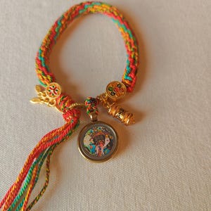May include: A colorful braided bracelet with a gold charm featuring a mandala design. The bracelet also has a gold pendant with a colorful image of two people and a gold charm with a small bell.