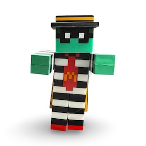 Puede incluir: Una figura en bloques con una cara verde, sombrero y máscara negros, vestida con un atuendo a rayas blanco y negro. Tiene brazos verdes en bloques y una corbata roja con el logo dorado de la "M". Se ve una capa amarilla.